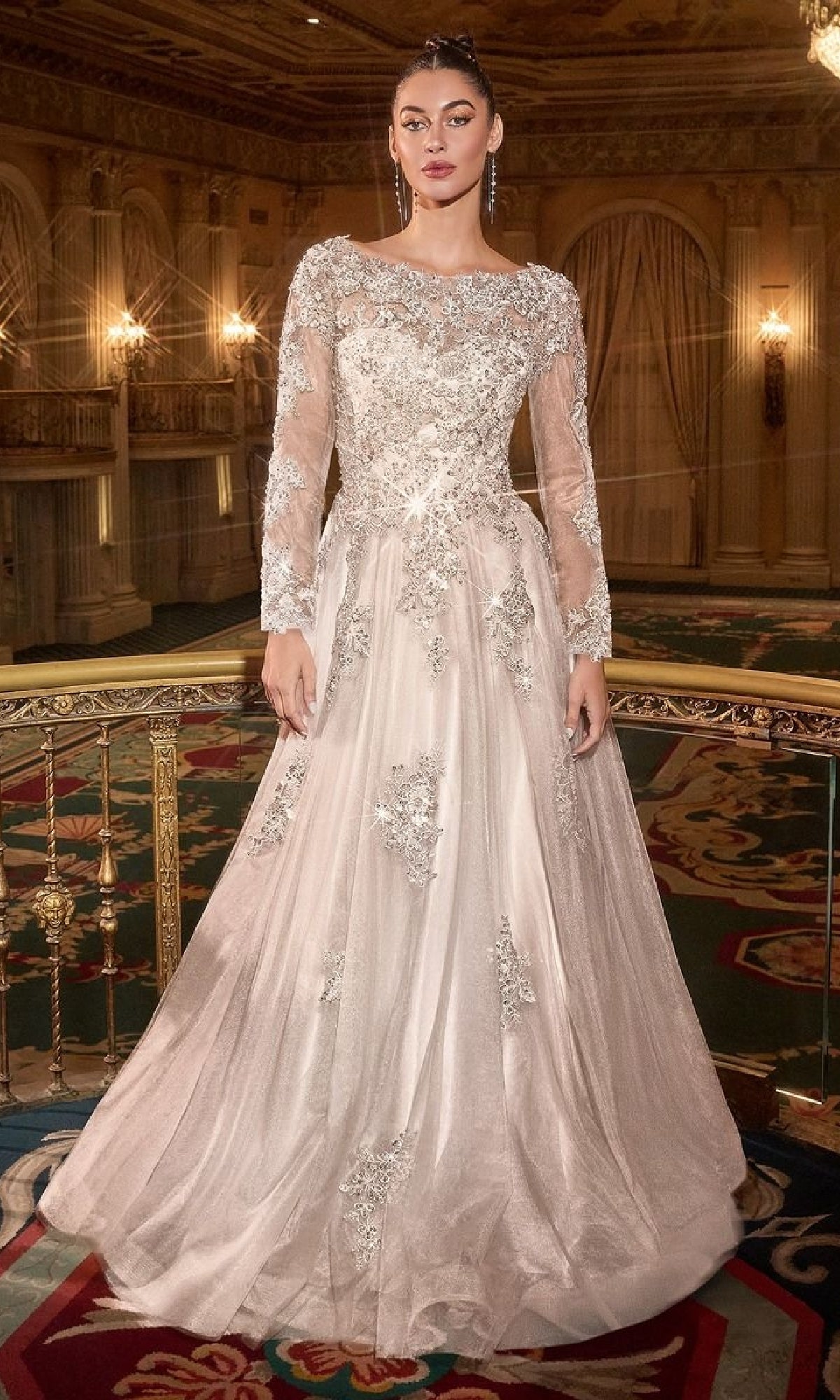 A woman stands in an ornate ballroom, wearing the Ladivine J883W—an elegant long sleeve formal dress with lace details and sheer sleeves, her light-colored gown radiating timeless sophistication.